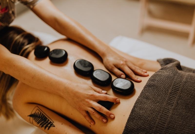 Una persona recibe un masaje con piedras calientes, tumbada boca abajo con seis piedras negras colocadas a lo largo de su espalda desnuda, mientras las manos de un terapeuta presionan suavemente las piedras. La persona tiene un tatuaje visible en el brazo izquierdo.