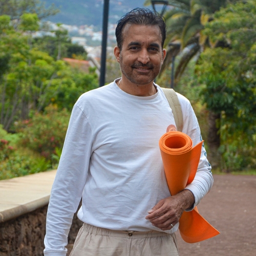 Un hombre vestido con camisa blanca de manga larga y pantalones claros sonríe mientras sujeta una esterilla de yoga naranja y una bandolera. Está al aire libre en un camino rodeado de vegetación y árboles.