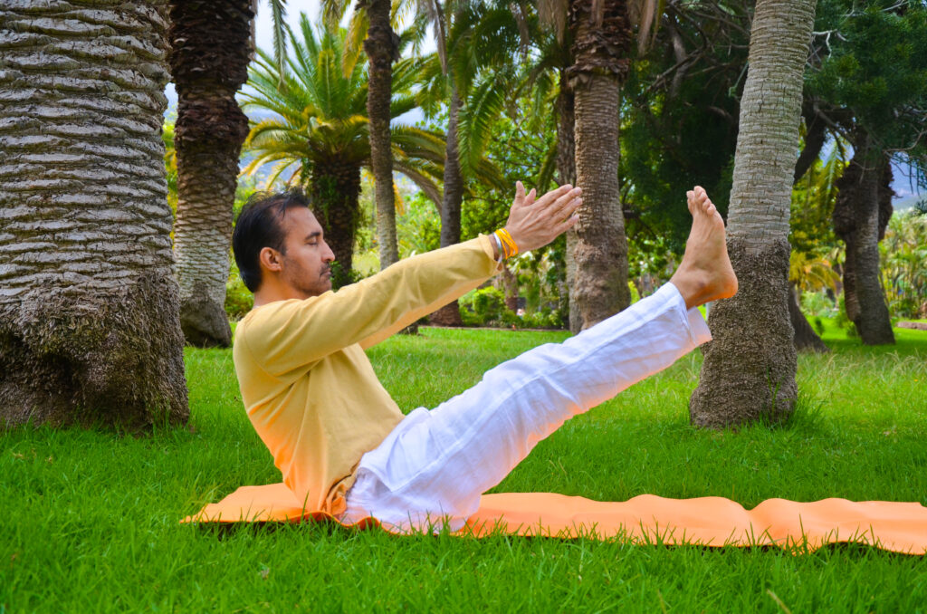 Un hombre practica yoga al aire libre sobre una esterilla, sentado en la hierba verde con las piernas y los brazos levantados, rodeado de palmeras en un parque exuberante y soleado.