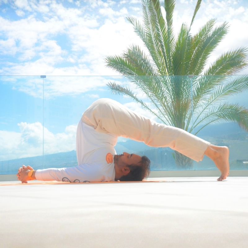 Una persona practica yoga al aire libre en la postura del arado, tumbada boca arriba con las piernas extendidas por encima de la cabeza y los pies tocando el suelo. Al fondo, una palmera, un cielo azul y una barandilla de cristal.