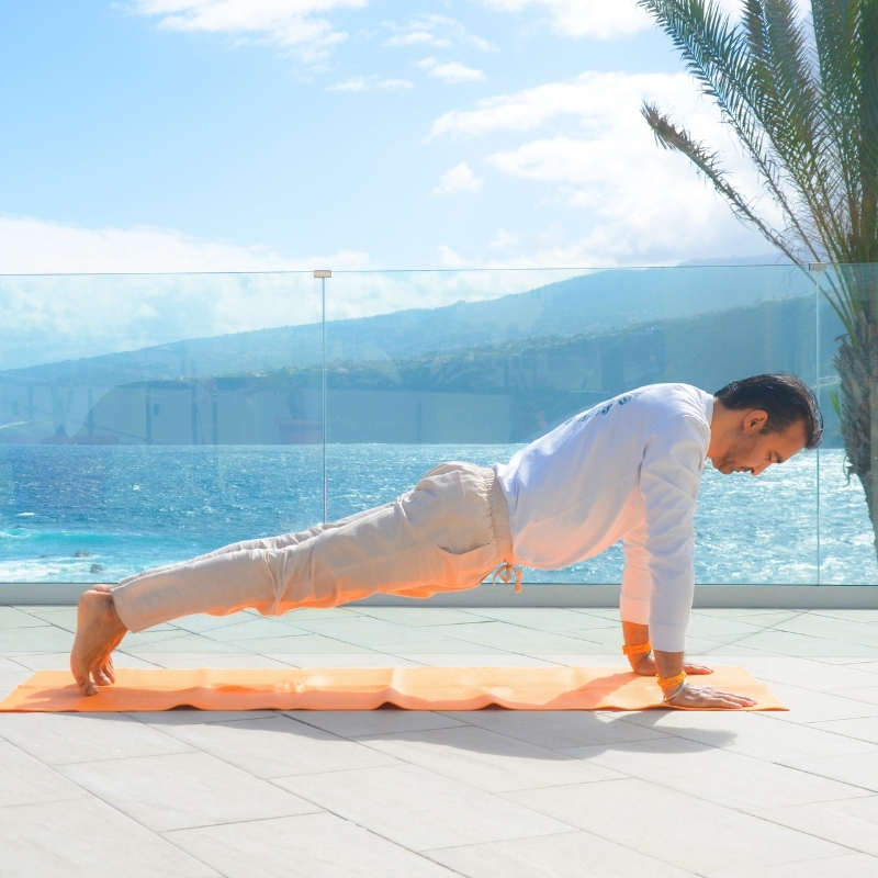 Una persona practica una postura de yoga en plancha sobre una esterilla naranja al aire libre, con vistas al océano, las montañas y las palmeras bajo un cielo parcialmente nublado.