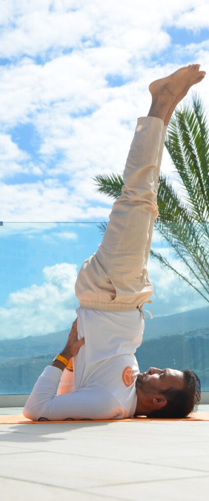 Un hombre practica una postura de yoga con los hombros al aire libre sobre una esterilla, con cielo azul, nubes, una palmera y montañas visibles al fondo.
