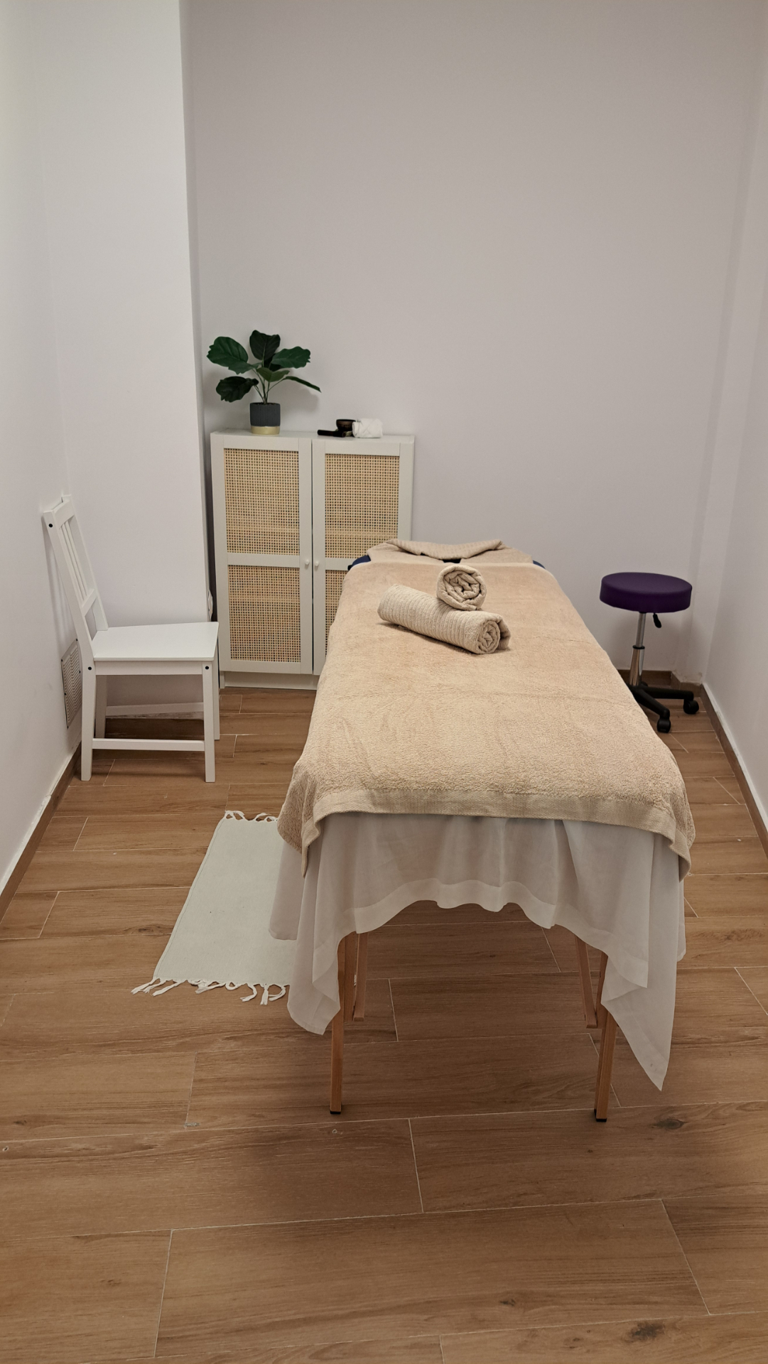 Sala de masaje con camilla, silla blanca, alfombra y pequeno taburete.
