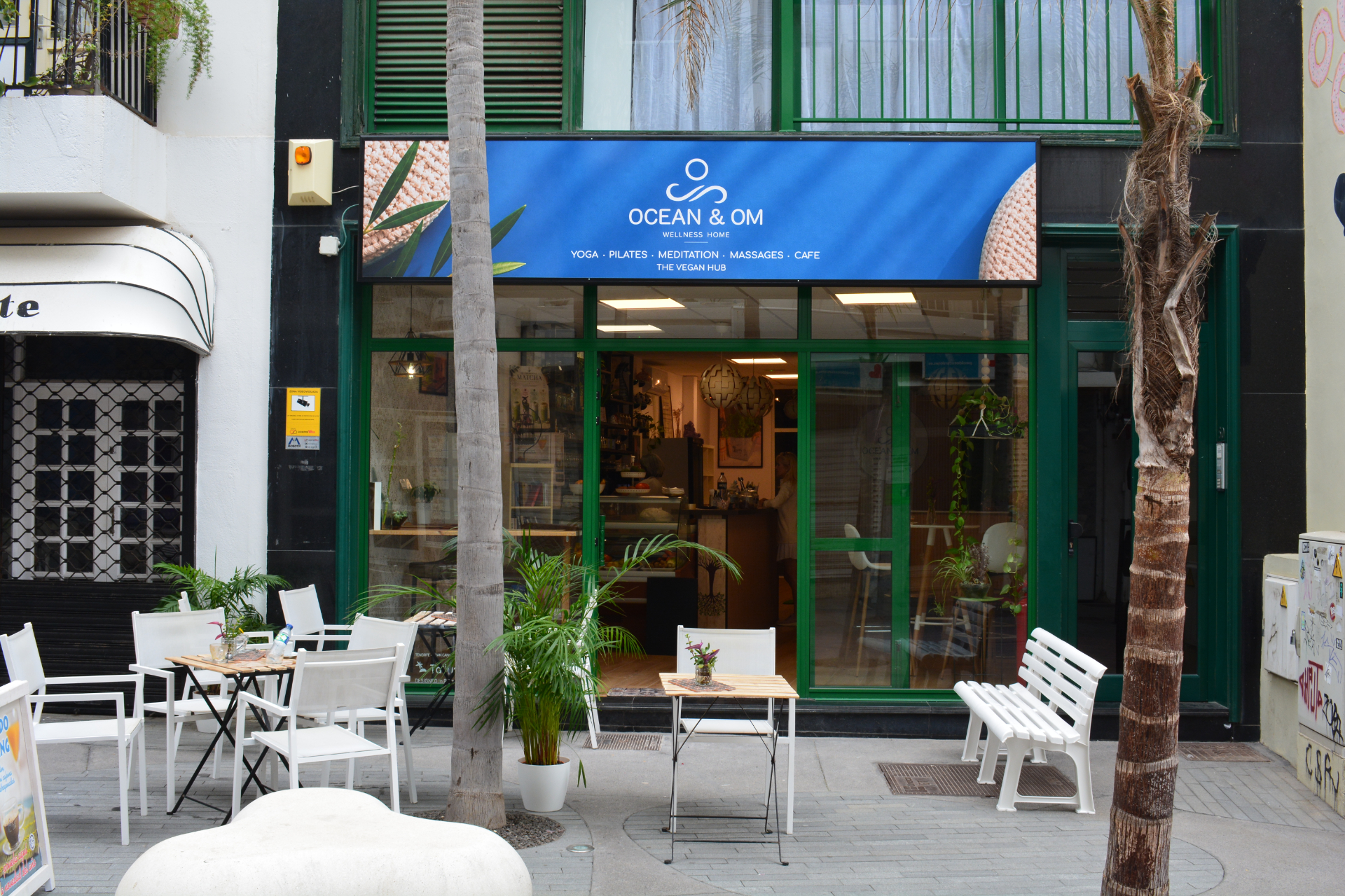 Street view of Ocean & Om, a yoga, pilates, and wellness centre with a café. The shopfront has large green-framed windows, outdoor tables and chairs, potted plants, and a blue sign with the business name.