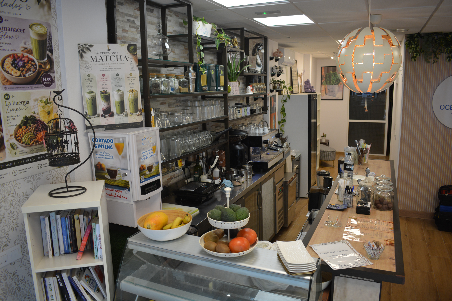 A cosy café interior with shelves holding glassware, tea, and plants. The counter displays fruit, desserts, and coffee equipment. Posters, books, and a round pendant lamp add to the welcoming atmosphere.