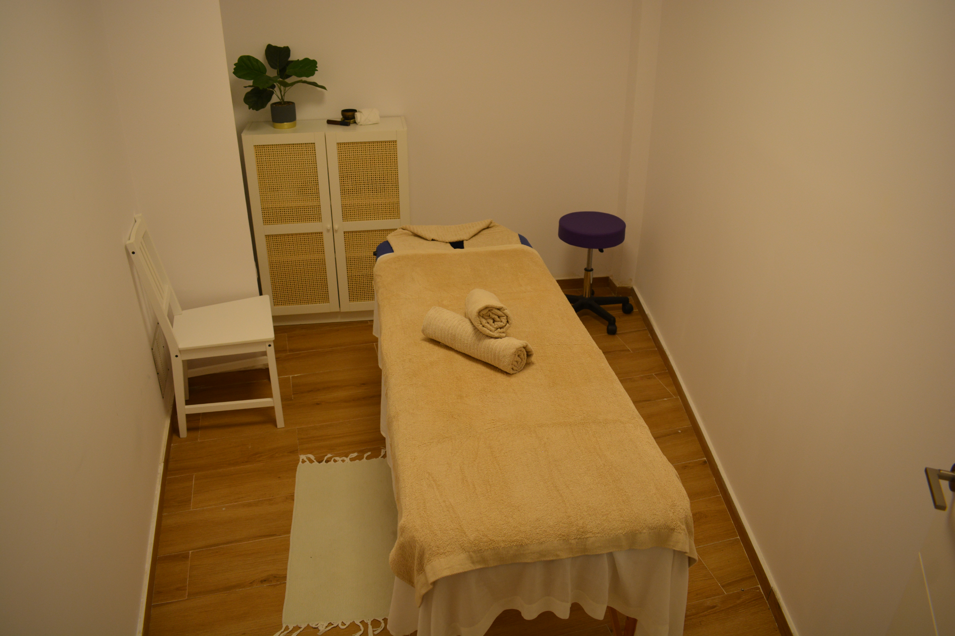 A small, neatly arranged massage room with a massage table covered in beige towels, a white chair, a purple stool, a cupboard with a plant on top, and wooden flooring.