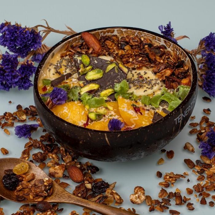 Un bol de batido con granola, rodajas de mango, pistachos, chocolate negro, semillas de chía y flores moradas comestibles, rodeado de granola esparcida, frutos secos y más flores moradas. Cerca hay una cuchara de madera.