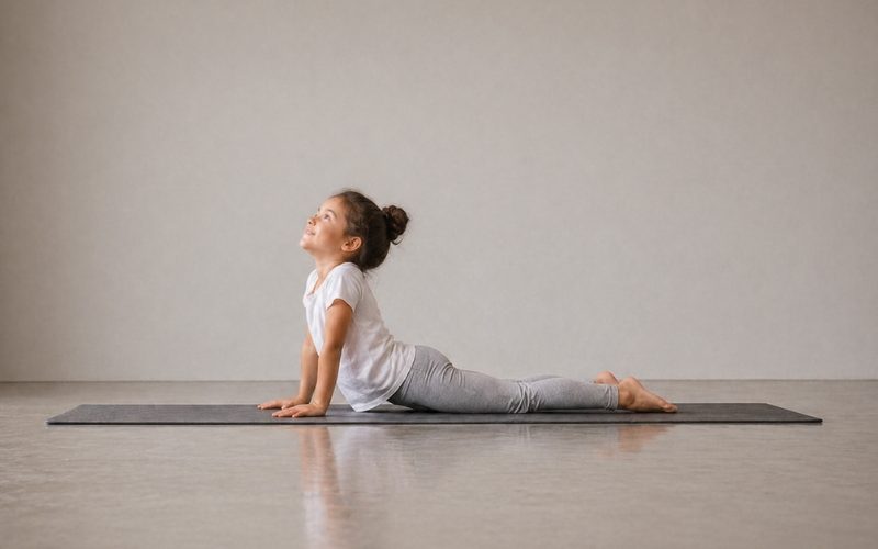 Una joven practica yoga en el interior sobre una esterilla, realizando la postura del perro mirando hacia arriba. Lleva una camiseta blanca y leggings grises, con el pelo recogido en un moño, mirando hacia arriba en una habitación luminosa y minimalista.
