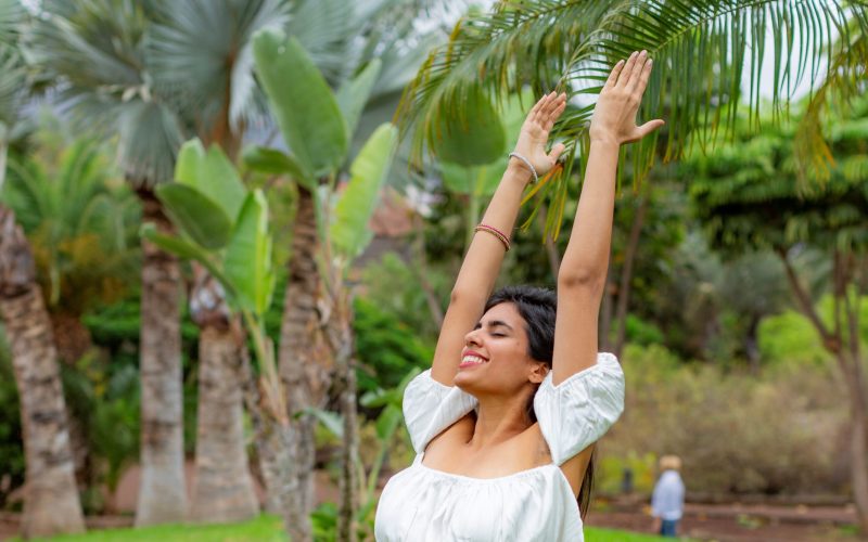 Una mujer con un top blanco está de pie al aire libre con los brazos levantados y los ojos cerrados, sonriendo alegremente. Está rodeada de exuberantes palmeras verdes y plantas en un jardín tropical.