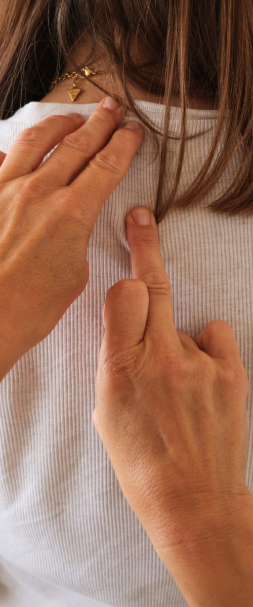 Una persona toca suavemente la parte superior de la espalda de otra con ambas manos, una mano en el cuello y la otra apuntando al centro de la espalda. La persona tocada lleva una camisa blanca.