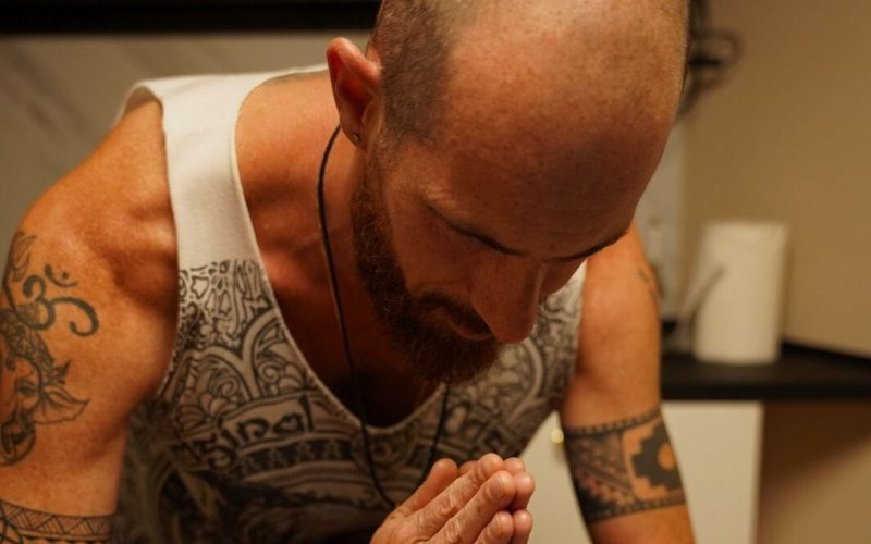 Un hombre calvo, con barba y tatuajes, inclina la cabeza y junta las manos sobre alguien tumbado, como si diera una bendición o se preparara para un masaje en una habitación suavemente iluminada.