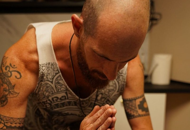 Un hombre calvo, con barba y tatuajes, inclina la cabeza y junta las manos sobre alguien tumbado, como si diera una bendición o se preparara para un masaje en una habitación suavemente iluminada.