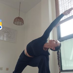 Una mujer con una coleta trenzada practica yoga en interiores, realizando una postura de ángulo lateral extendido sobre una esterilla de yoga. Estira un brazo por encima de la cabeza mientras dobla la rodilla delantera, en una habitación luminosa con grandes ventanas y luz natural.