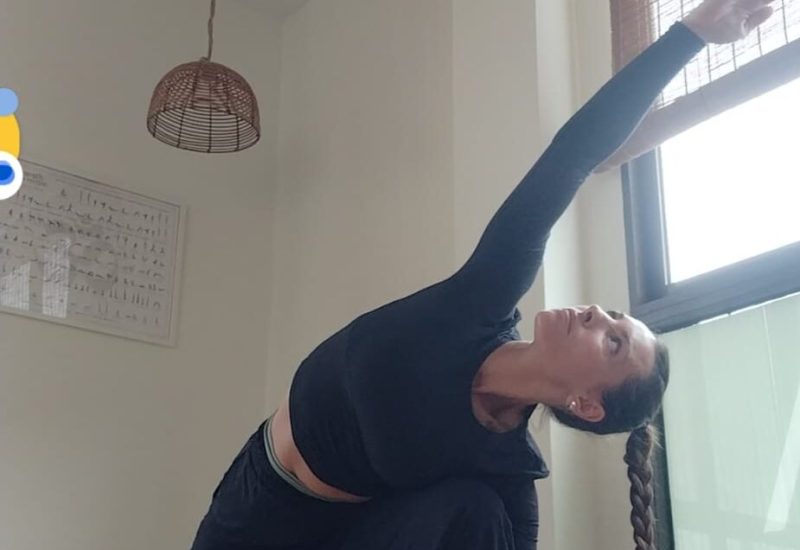 Una mujer con una coleta trenzada practica yoga en interiores, realizando una postura de ángulo lateral extendido sobre una esterilla de yoga. Estira un brazo por encima de la cabeza mientras dobla la rodilla delantera, en una habitación luminosa con grandes ventanas y luz natural.