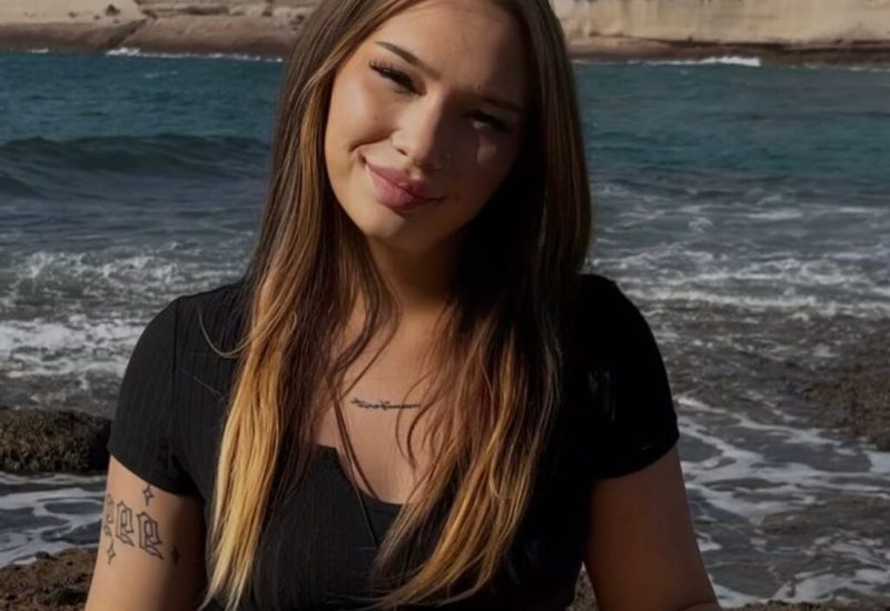 Una mujer con el pelo largo y castaño y tatuajes en el antebrazo sonríe a la cámara mientras está sentada sobre unas rocas junto al mar, con acantilados y olas de fondo en un día soleado.